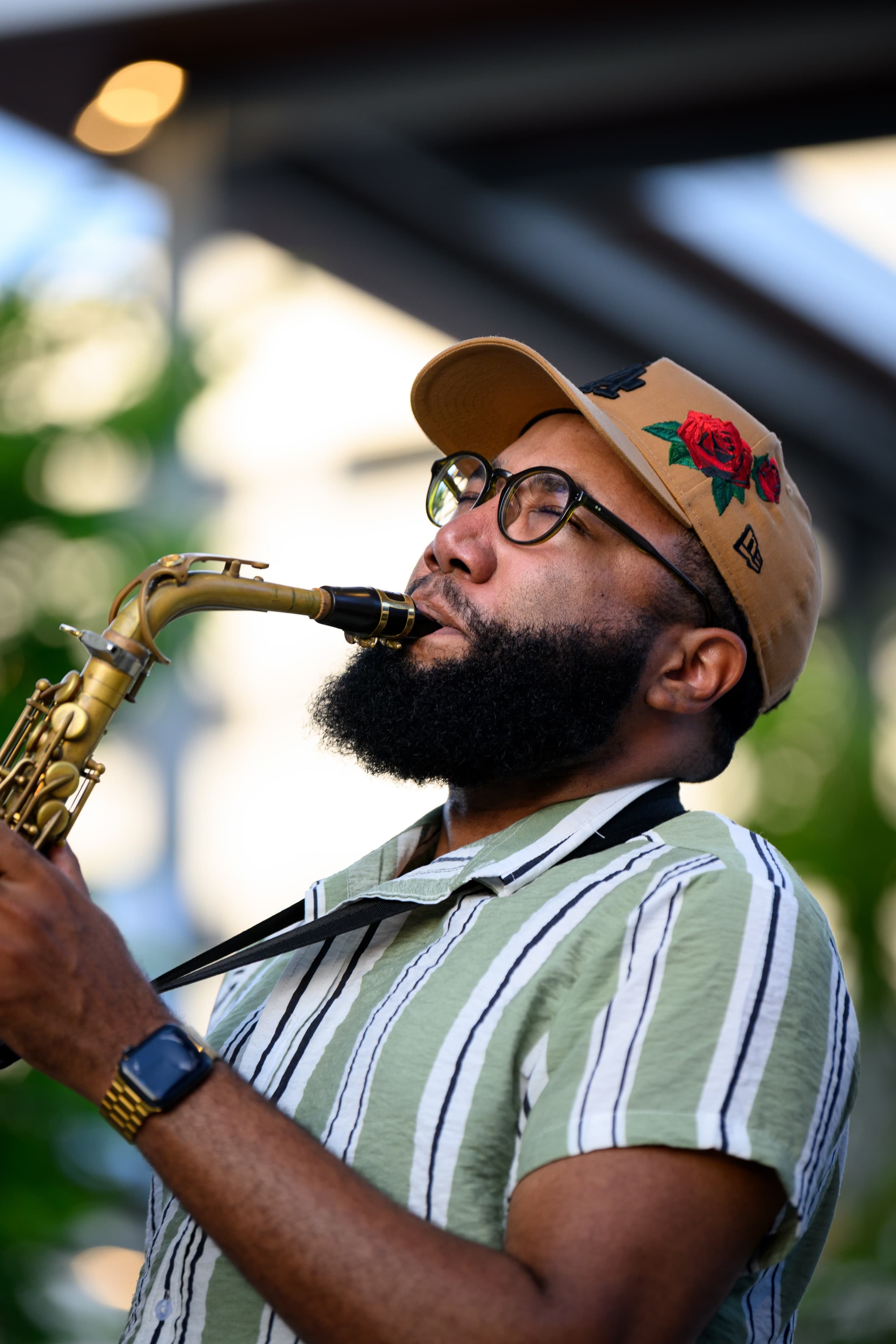 Bearded man wearing glasses and a rose-embroidered baseball cap playing a saxophone outdoors.