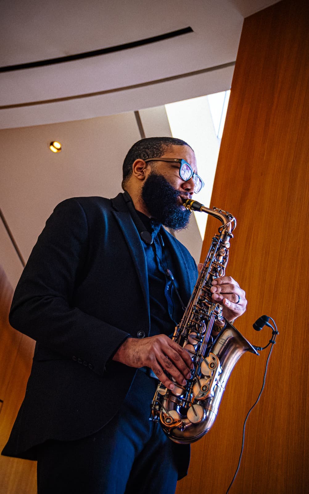 Bearded Black man in a black suit playing a saxophone with a clip-on microphone.