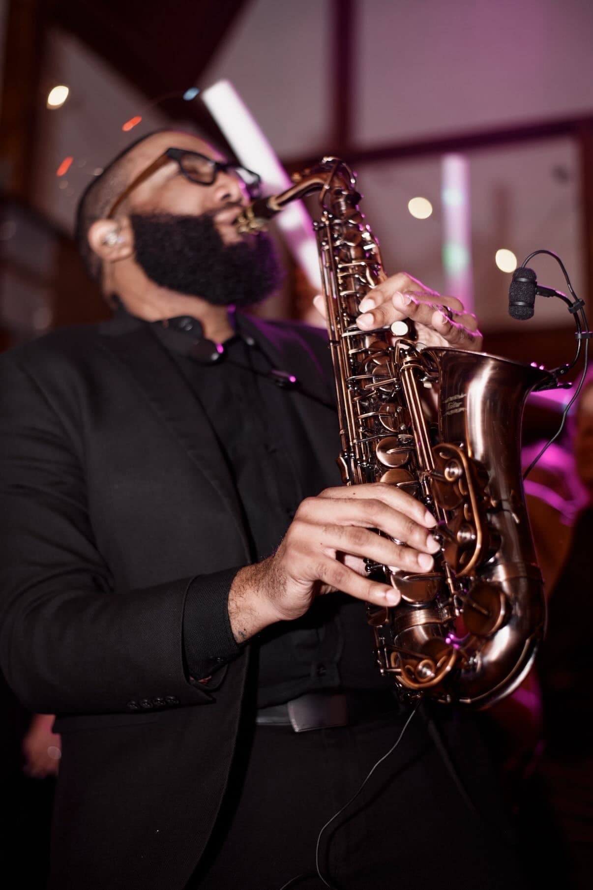 Bearded musician in a black suit plays a bronze saxophone under purple stage lighting.