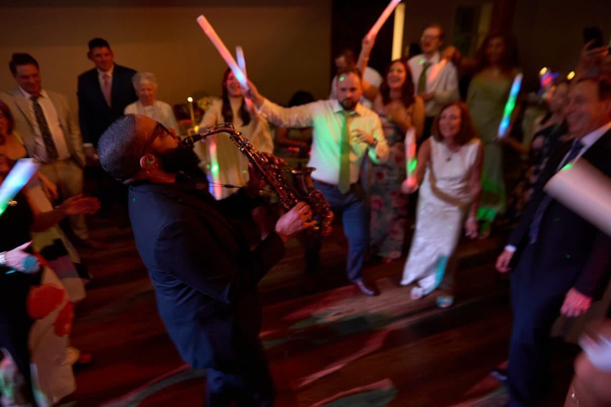 A saxophonist performs while guests dance with glowing foam sticks at a lively celebration.