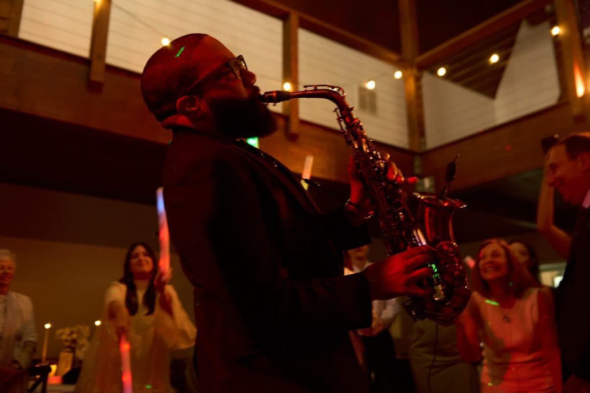 Bearded man plays saxophone at a dimly lit indoor event with guests dancing behind him.