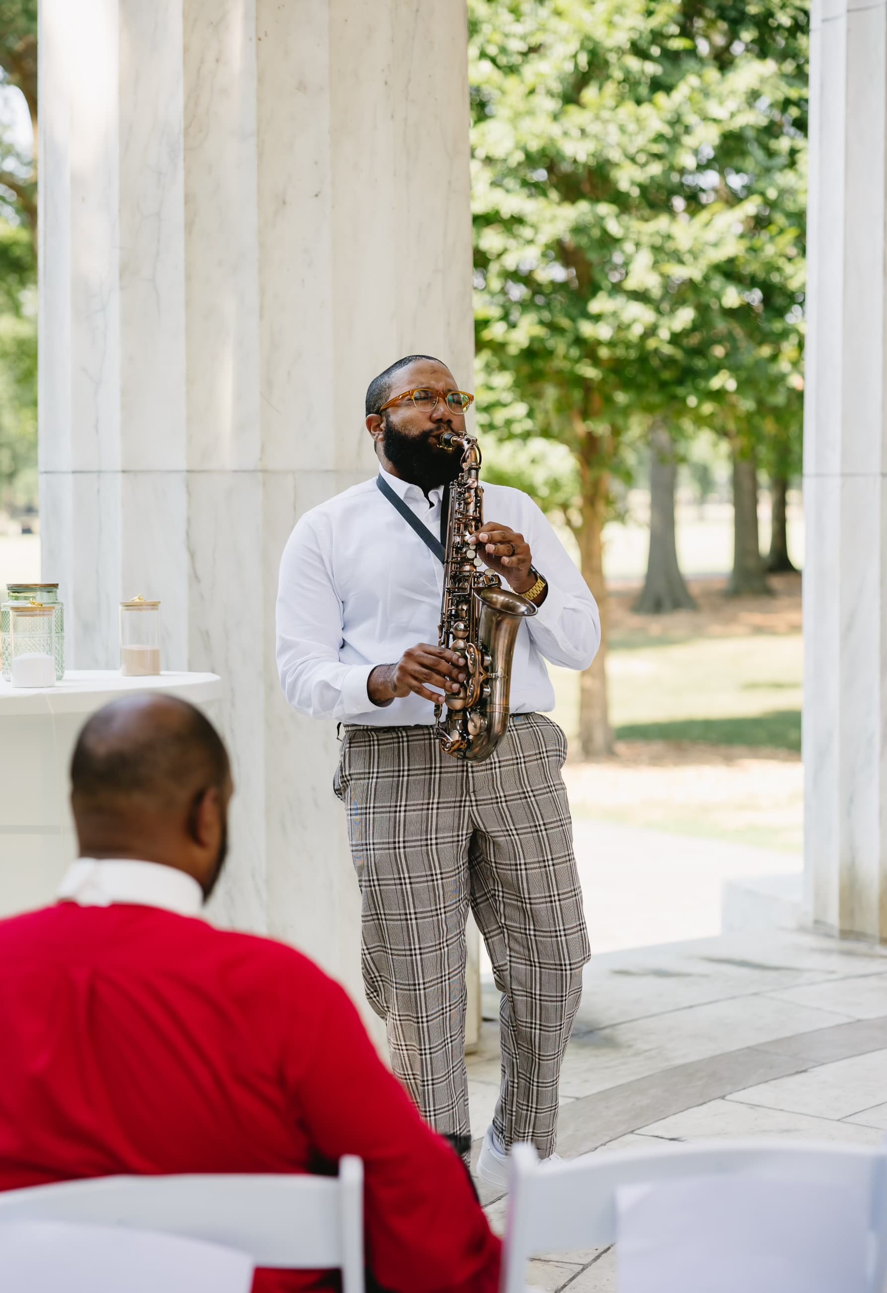Bearded man in white shirt and plaid trousers plays saxophone outdoors near white marble columns.