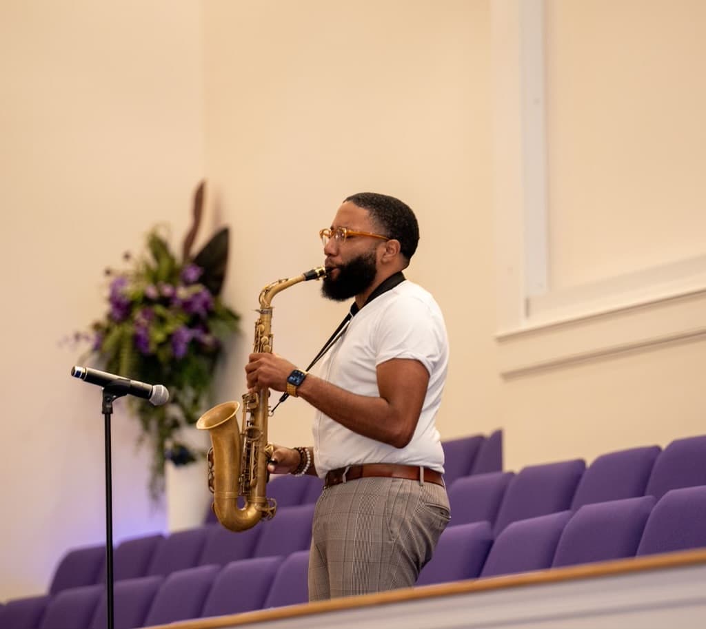 Bearded man in a white shirt plays a saxophone before rows of purple chairs.