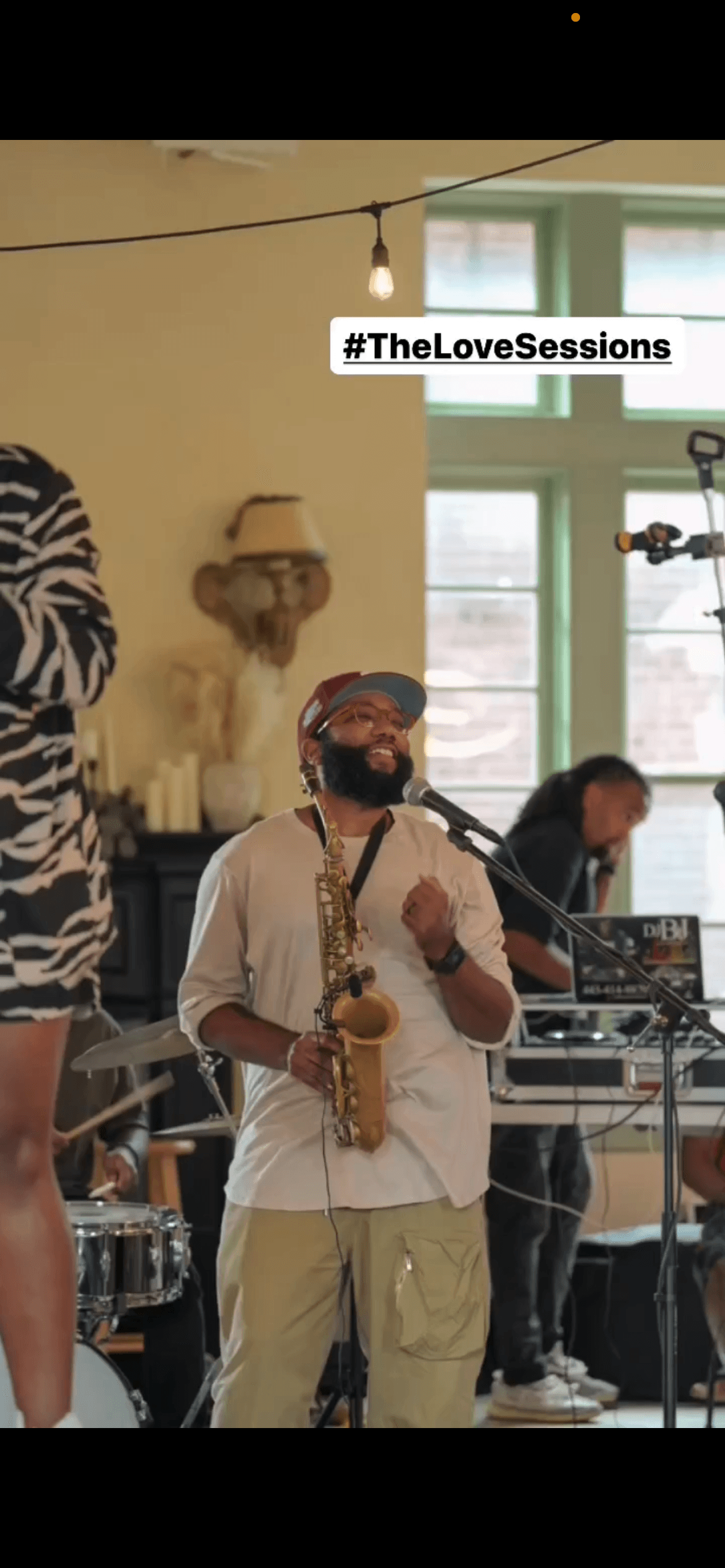 Smiling bearded man in a red cap holds a saxophone during a live music session.