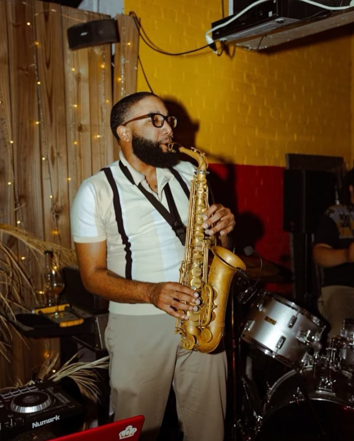 Bearded man in glasses playing a saxophone in front of a vibrant yellow wall.