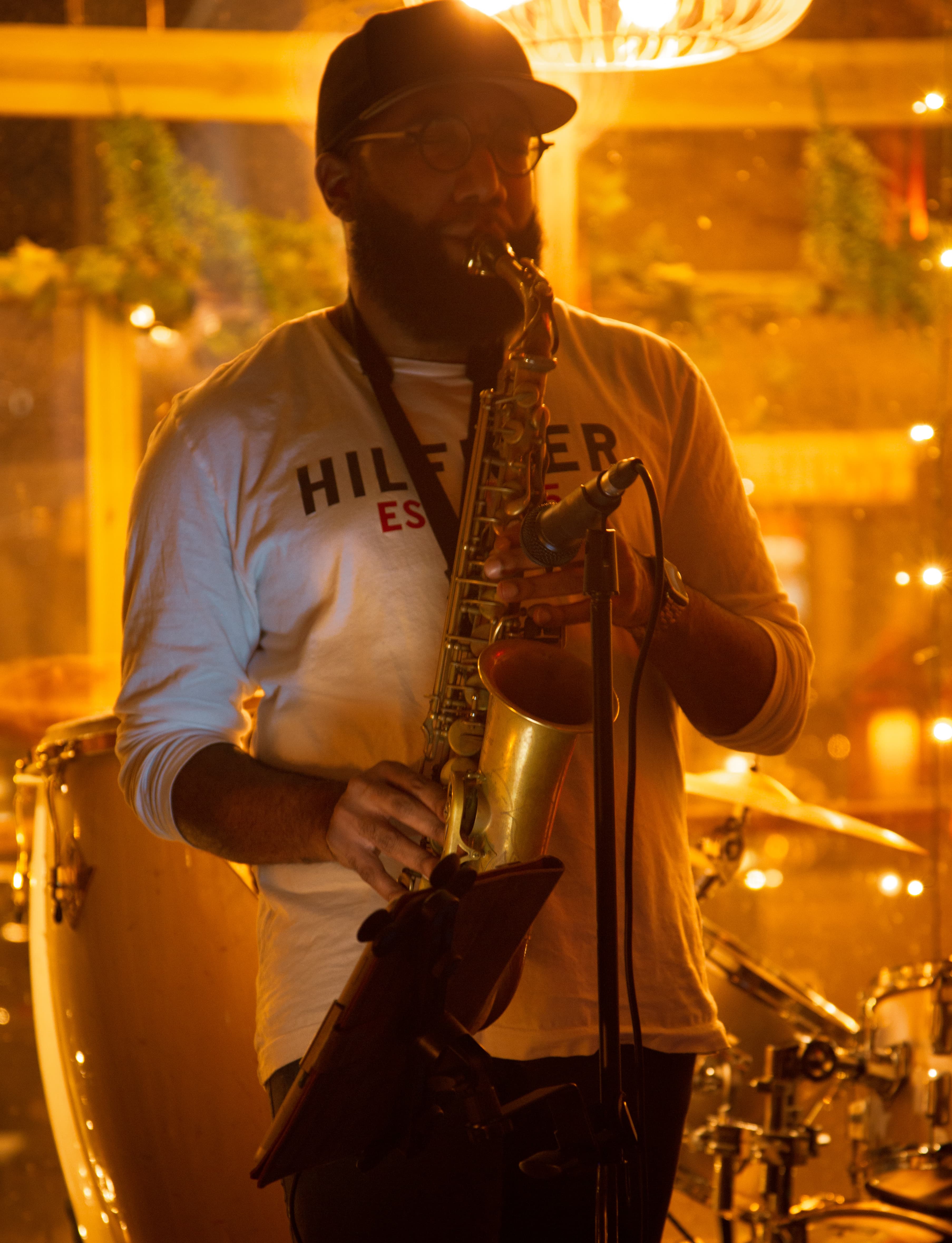 Bearded man in a cap plays saxophone under warm, golden lighting at a live performance.