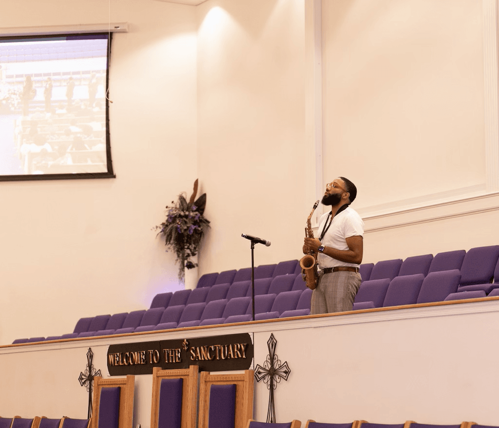 Bearded man playing saxophone in a church sanctuary with purple seats and a welcome sign.